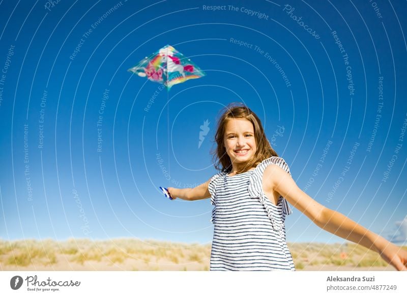 Cute happy little girl in summer dress running with flying kite on empty sandy beach. Beautiful sunny day, blue sky. active activity beautiful carefree cheerful