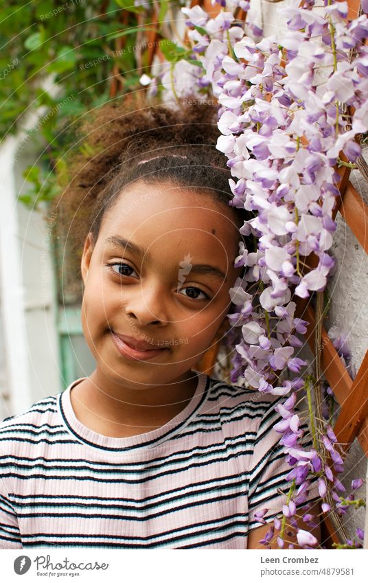 Teenage girl of mixed race with curly hair posing in between purple wisteria flowers Girl teen teenager beauty curls summer green plant portrait outside emotion