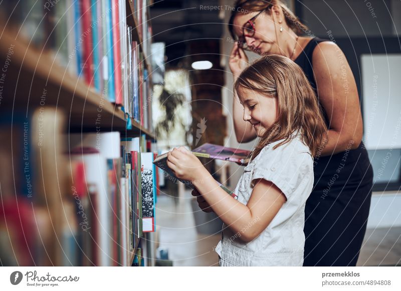 Teacher helping to choose book her schoolgirl in school library. Smart girl selecting literature for reading. Books on shelves in bookstore. School education. Benefits of everyday reading