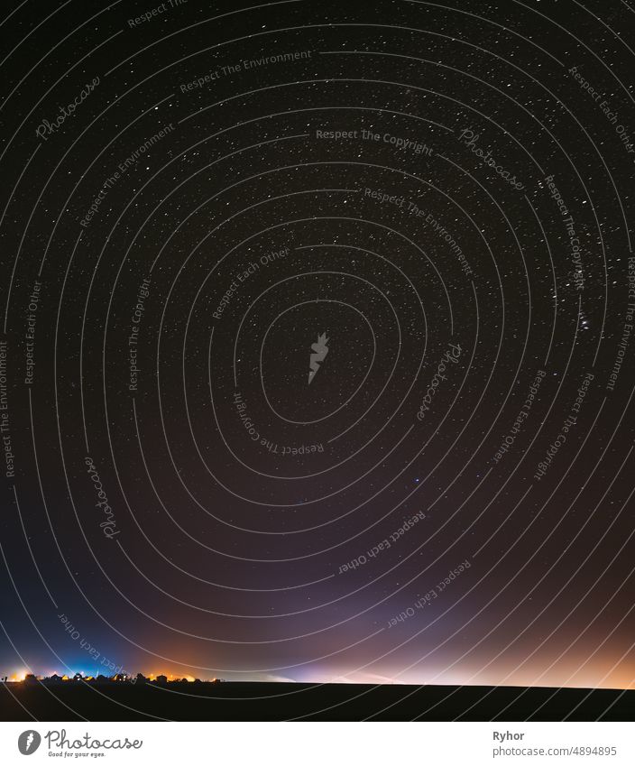 Night Starry Sky With Glowing Stars Above Countryside Town Landscape. Rural Field Meadow In Early Spring. Bright Glow Of Stars. Illumination Lights Of Small Town Or Village. Milky Way Galaxy