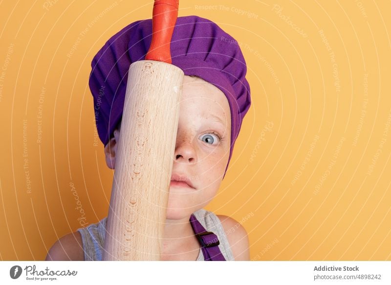 Cute little child holding rolling pin near face and looking at camera boy chef funny playful cover eye kitchenware portrait grimace cook childhood culinary