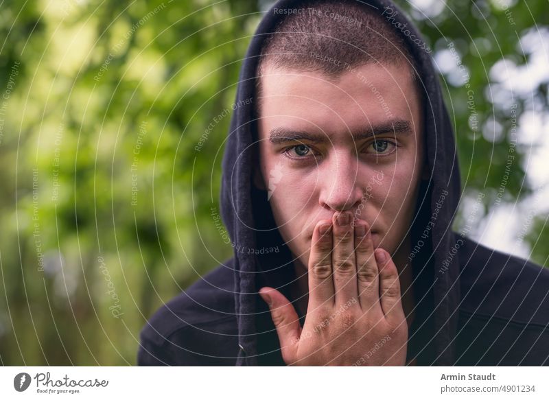 portrait of young serious man wearing hoodie and hand over mouth covering silent finger look teenager looking male beautiful casual caucasian outdoor confident