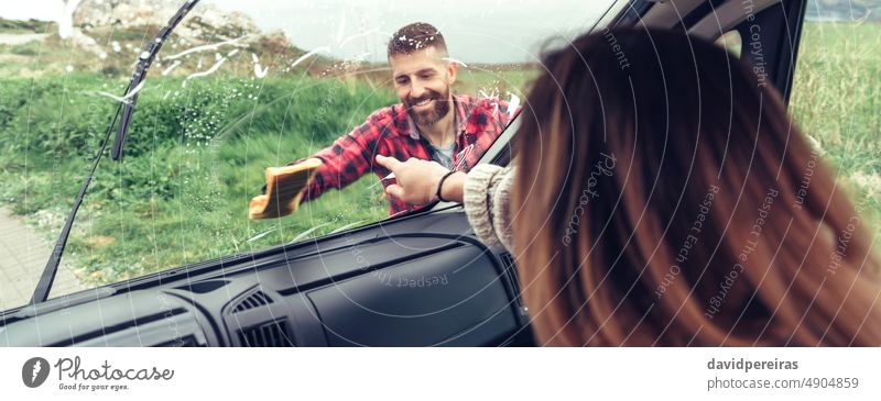 Man cleaning van windshield while woman pointing stain smiling rinsing glass banner web header panorama panoramic cloth copy space view from inside