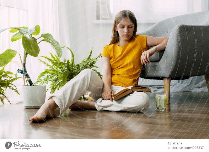 A young female student is sitting on the floor and reading a book, enjoying reading on a weekend or preparing for classes at school or university. Back to school, preparing for classes, free time