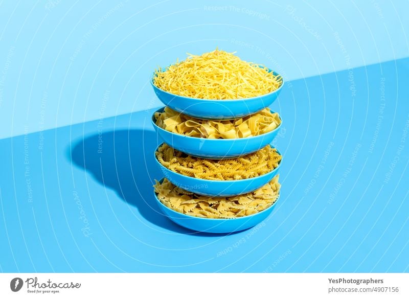 Raw pasta variety on a blue table. Stack of plates with uncooked pasta assortment background bowls bright carbs color cuisine different dry farfalle fettuccine