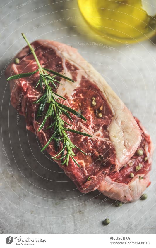 Close up of raw beef steak with rosemary and olive oil at grey concrete kitchen table. close up butcher background fresh beef meat top view barbecue cooking