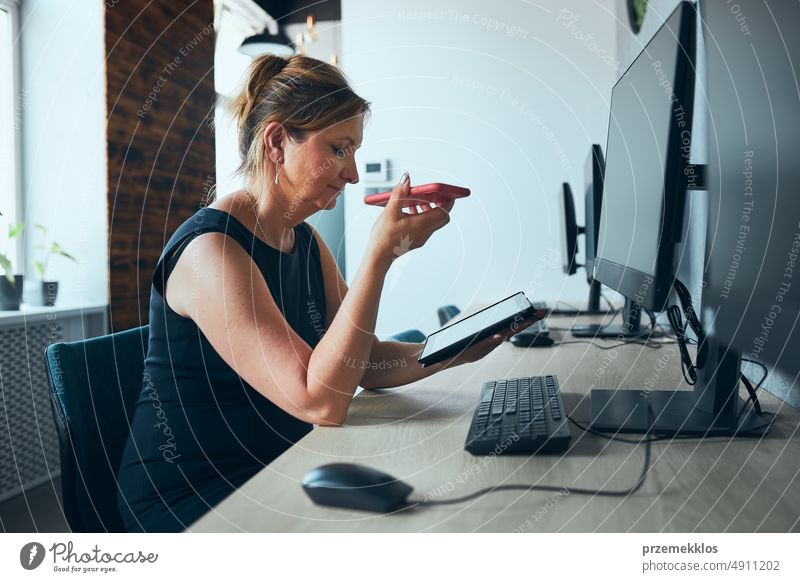 Businesswoman having business call working on tablet in office. Mature busy woman using touch pad computer standing by window in modern interior smartphone talk
