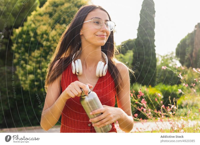 Content woman with water bottle in a park headphones refreshment beverage forest city female town lady summer casual young outdoors brunette long hair walking