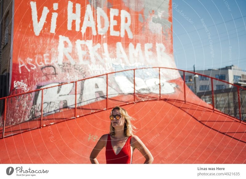 It’s hot in the city of Copenhagen, Denmark. A smoking hot blonde girl dressed in a red swimsuit is standing all proud behind a red skatepark with some graffiti and tags on it. Summer in the city might be adventurous as well.