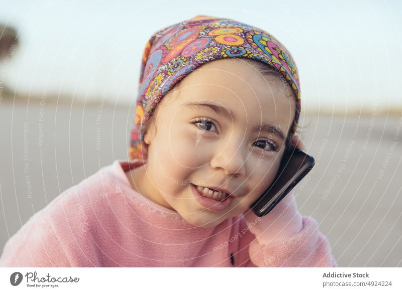 Happy child sitting on road and speaking on smartphone girl happy weekend asphalt smile daytime cloudy sky hispanic ethnic preschooler casual headscarf kid