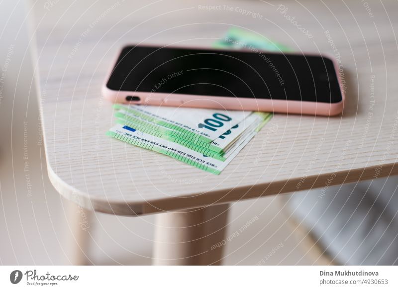 Mobile phone on a pile of green 100 (one hundred) euro banknotes closeup. Inflation in European Union concept. Money and technology. Pile of one hundred euro notes on wooden table at home. Cash payments concept.