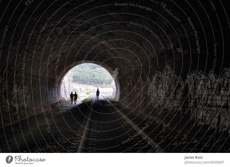 the exit of an old dark railroad tunnel seen from the inside with the silhouette of three people walking in the distance coming out of the tunnel, concept of overcoming