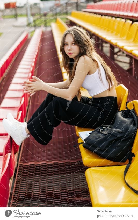 A smiling teenage girl is sitting on the podium next to her school backpack bleachers stadium student studying university high school resting near young female