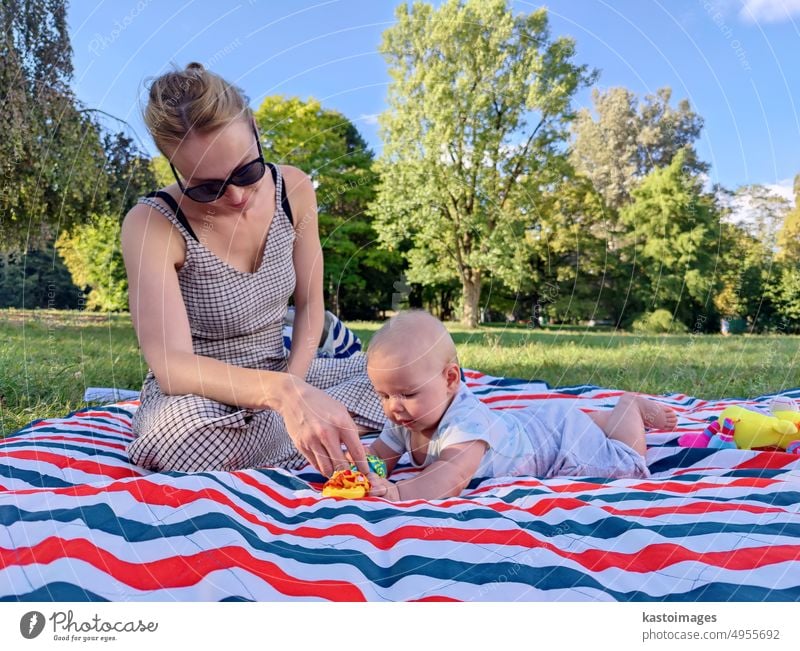 Modern trendy mum playing with his baby boy child on a mat in a meadow outdoors in public city park motherhood childhood woman lifestyle mom family breast happy