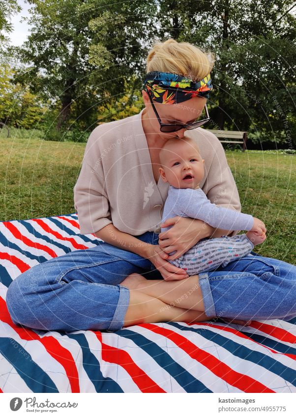 Modern trendy mum kissing and caressing his baby boy child on a mat in a meadow outdoors in public city park motherhood childhood woman lifestyle mom family