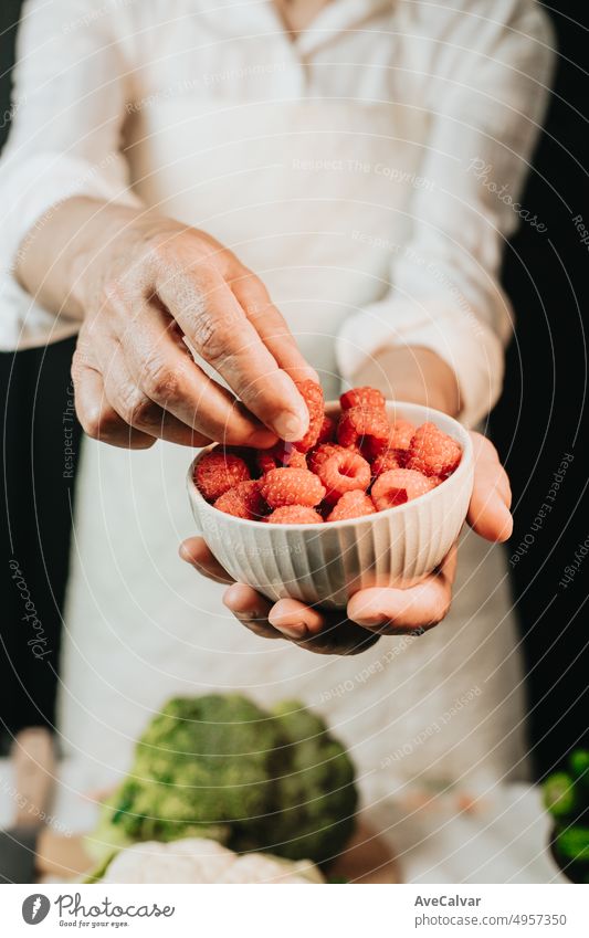 Woman holding a cup with raspberries and showing the quality in close up.Preparing the ingredients for a sauce to improve the recipe.Black background with white clothes and broccoli on the table