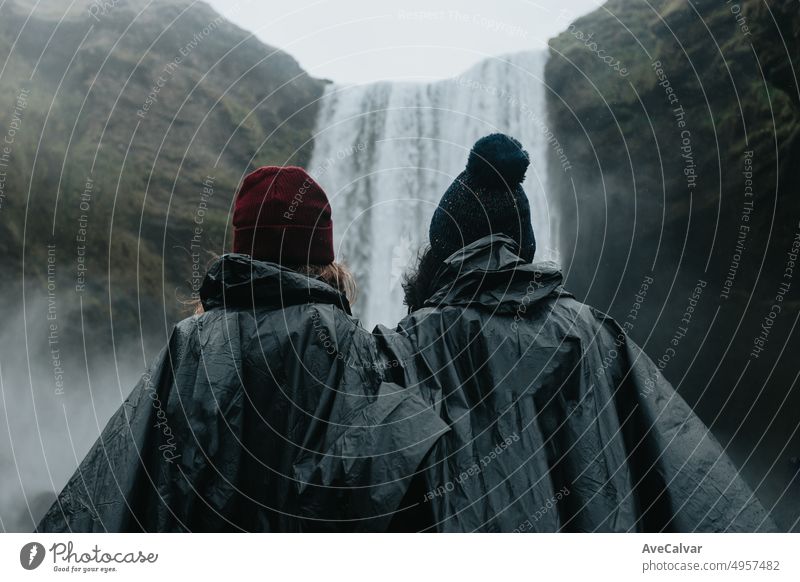 Woman couple in rain clothes in front of Skógafoss waterfall in Iceland during a moody day filled with water. Live your dream, love in Iceland, road trip style. Visit Iceland.Copy space image