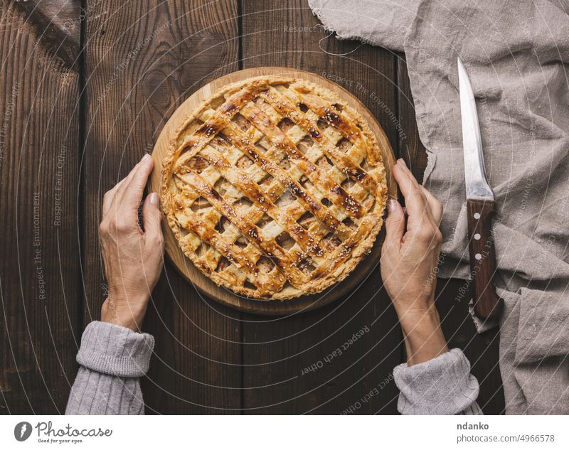 Two female hands hold a round baked pie with apple filling on a wooden board, brown table. dessert fruit food sweet homemade tart crust pastry fresh delicious