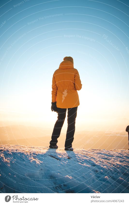 Traveller in a distinctive yellow jacket stands on top of a hill in the Polish Beskydy Mountains at sunset, enjoying the moment. Real people in winter ice environment