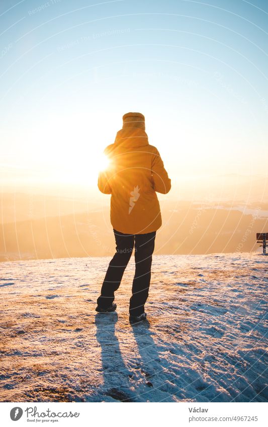 Traveller in a distinctive yellow jacket stands on top of a hill in the Polish Beskydy Mountains at sunset, enjoying the moment. Real people in winter ice environment