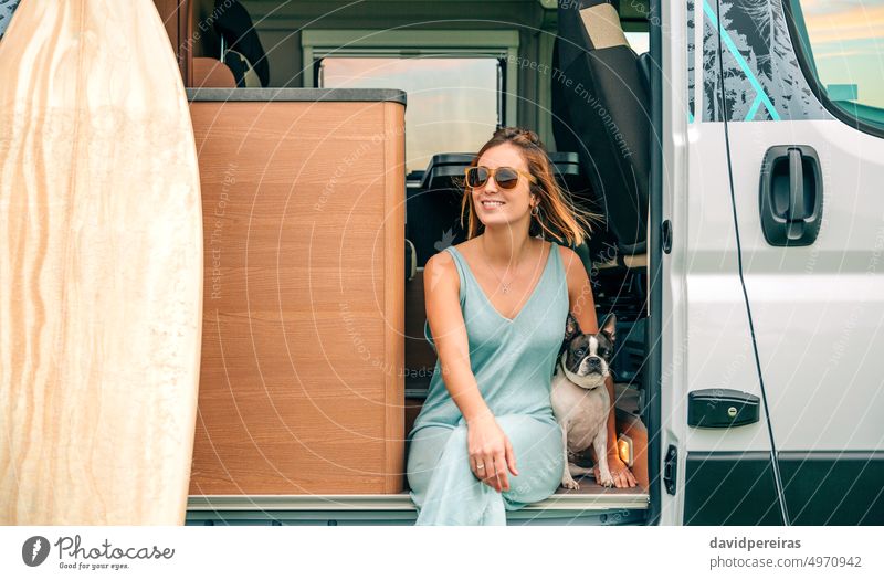 Woman sitting with her dog at the door of her camper van during a trip happy young woman smiling boston terrier looking aside laughing pet vacation breeze