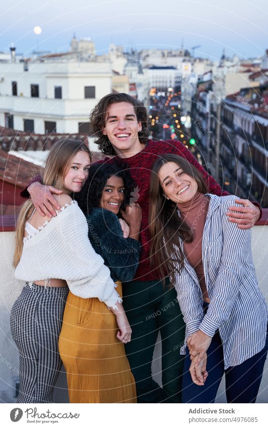 Smiling multiracial friends hugging and sitting on rooftop in twilight toothy smile cheerful together embrace carefree gather young terrace veranda town city