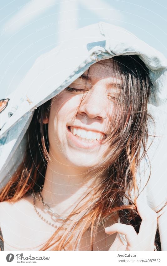 Super close up portrait of a happy young woman eyes closed covering head with jacket sunny day 20s smiling chatting friendly journey joyful laughing listen look