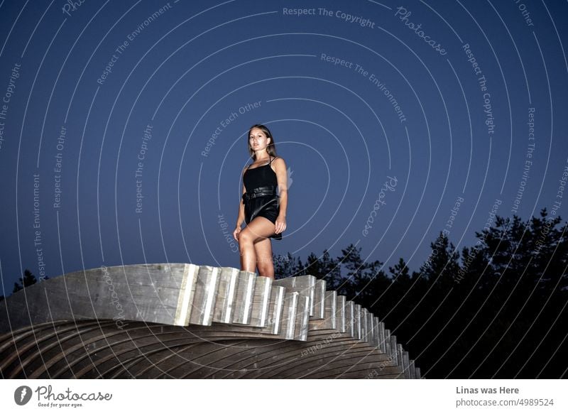 The sky is dark blue. A gorgeous brunette girl is dressed in black. Looking straight into a camera. She’s tanned and fashionable. A pretty woman and her model test in front of you.