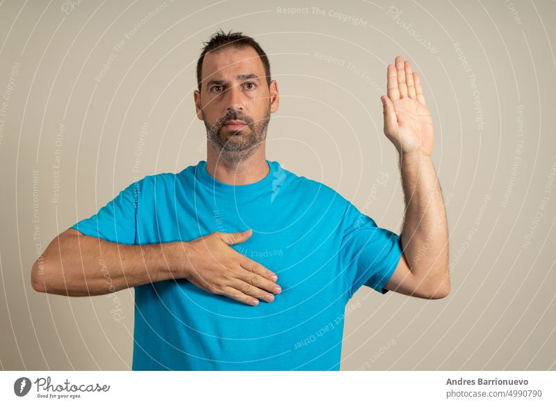 Bearded hispanic man in his 40s wearing blue t-shirt over beige isolated background swearing with hand on chest and open palm, making allegiance promise oath.