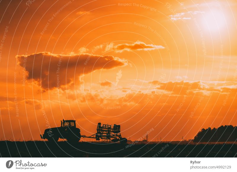Tractor Rides On Countryside Road. Beginning Of Agricultural Spring Season. Cultivator Pulled By A Tractor In Rural Field Landscape Under Sunny Summer Sunset Sunrise Sky. Backlit Dramatic Lighting