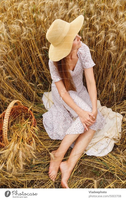 Young barefoot woman in white dress and hat sit in cereal field sun nature summer basket view wheat rye hair blonde wicker rural rustic country alone freedom