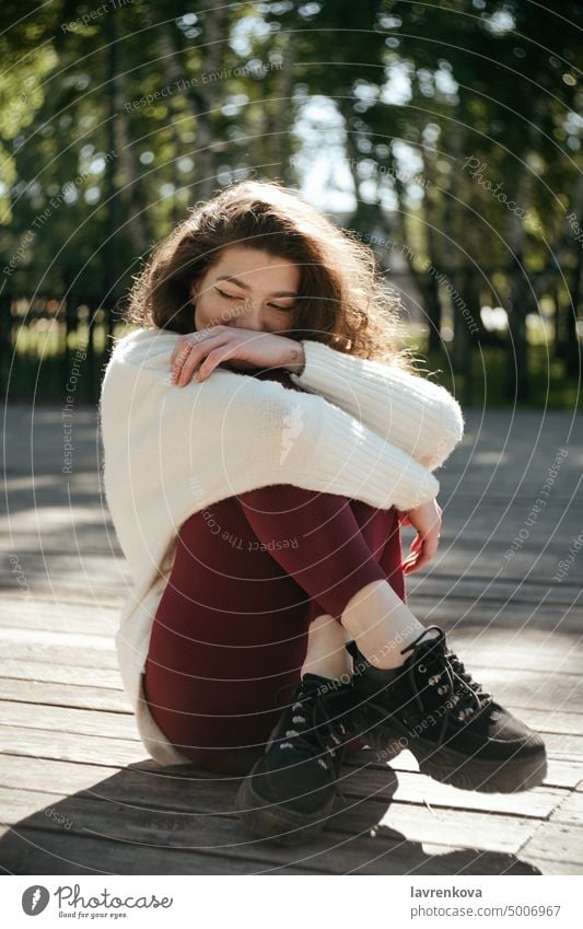 Smiling brunette woman in a white sweater sitting in the park, selective focus adult alone attractive autumn casual caucasian cheerful curly hair cute fall