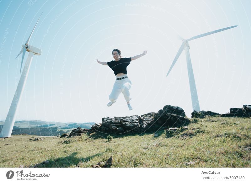 Young woman jumping and having fun celebrating freedom raising arms to the sky while smiling to camera surrounded by electric windmills. Blowing hair and relax, clean earth future concept