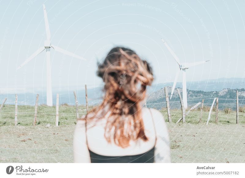 From behind young woman standing looking to surrounded by electric windmills. Blowing hair and relax, clean earth future concept person freedom turbine