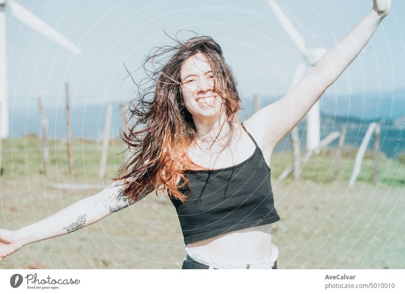 Frontal portrait happy young woman enjoying freedom raising arms to the sky while smiling to camera surrounded by electric windmills. Blowing hair and relax, clean earth future concept