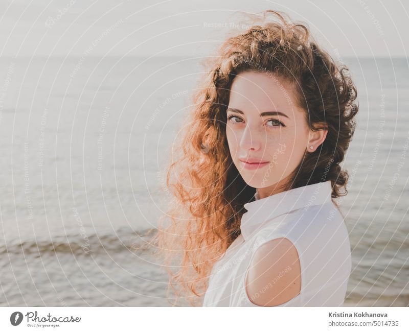 Beautiful girl with curly long hair towards the camera on sunny spring sea beach. Young stylish business woman in fashion black coat and white shirt near ocean.