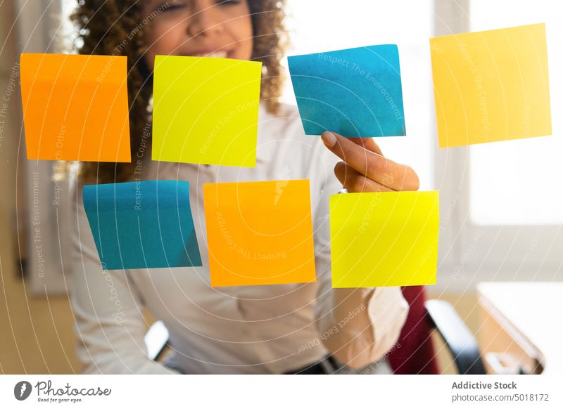 Crop smiling ethnic employee putting paper stickers on transparent wall worker sticky note data analyze strategy project startup smile woman office empty