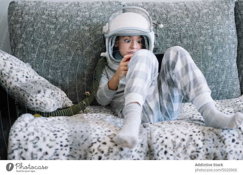 Kid lying on couch and playing with a video game console at home astronaut background boy child childhood concept costume creation handmade helmet imagination