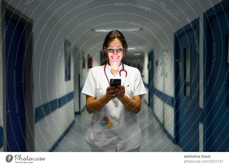 Cheerful pediatrician using smartphone in hospital hallway corridor walk smile uniform woman break doctor work job device gadget dark shift watching surfing