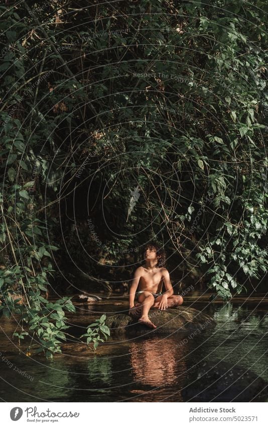 Dreamy boy resting on stone on river shore in forest dreamy trunks relax positive kid coast lush woodland nature brook calm stream flow woods child water