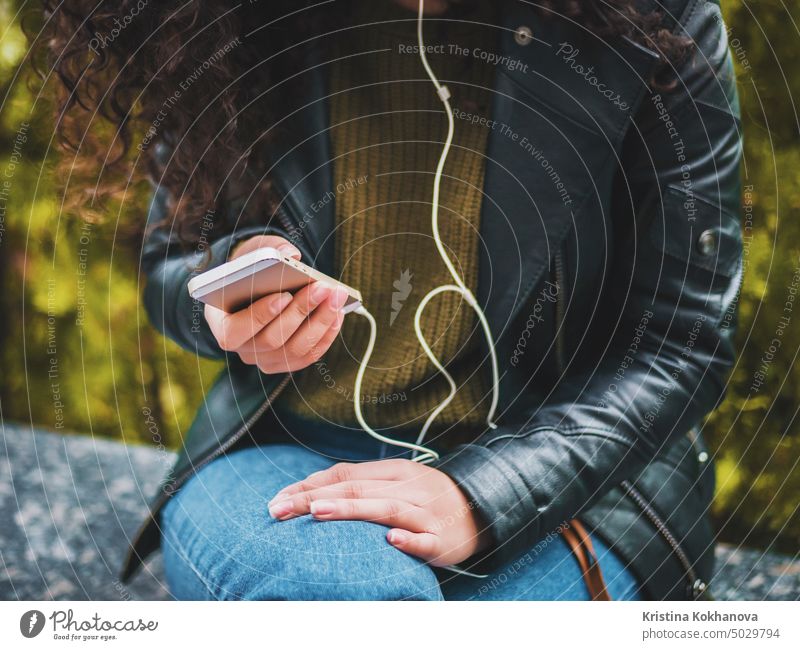 Pretty young latin girl with black curly long hair listening music with earphones and smartphone or player on a deserted autumn street. Woman enjoying moment.