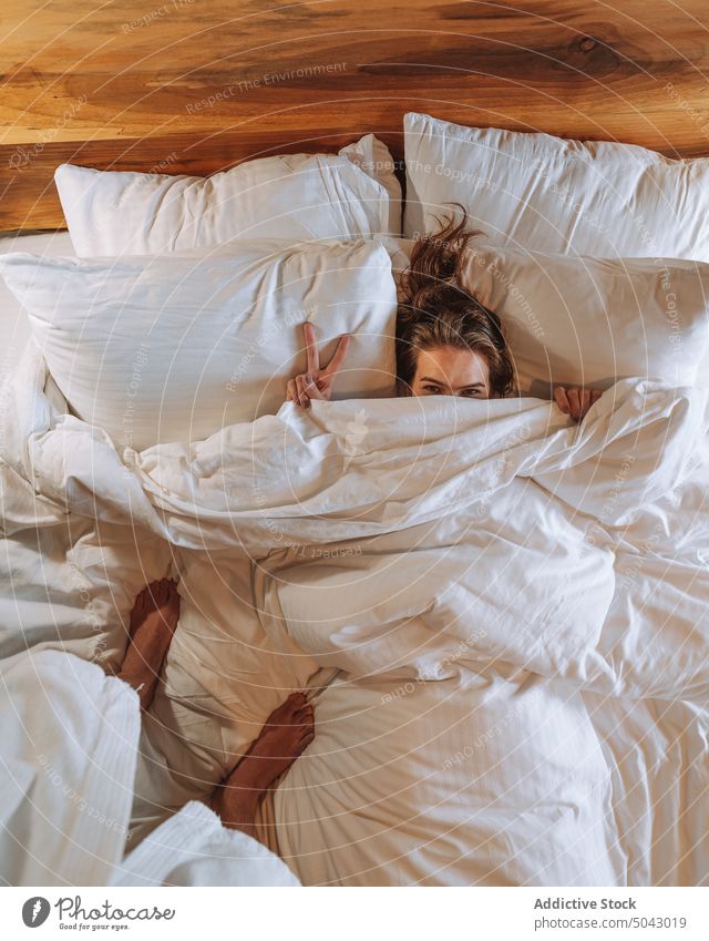 Happy woman showing V sign to crop boyfriend standing on bed couple v sign hotel romantic take photo positive together relationship lying hide relax bedroom