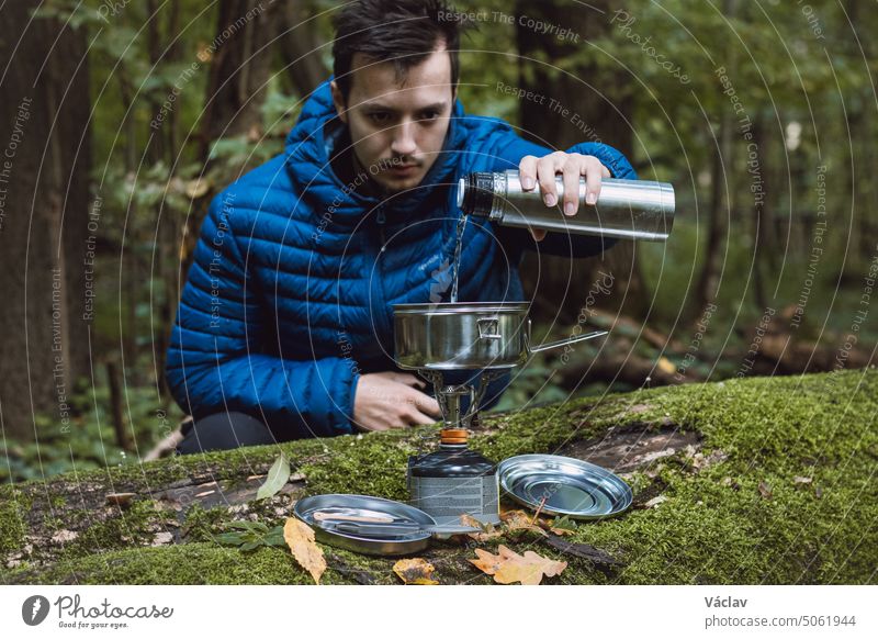Man in a blue down jacket boils water for tea and food on a portable stove and in a stainless steel ultralight pot in the middle of the wilderness. Hiking lifestyle