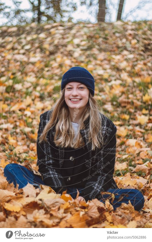 Smiling brunette sitting in a pile of colourful leaves in an oak forest throwing leaves. Candid portrait of a young real woman in autumn clothes in the autumn season