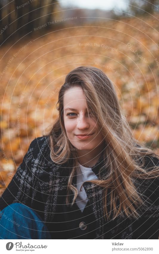 Smiling brunette sitting in a pile of colorful leaves in an oak forest. Candid portrait of a young realistic woman in autumn clothes in the fall season girl