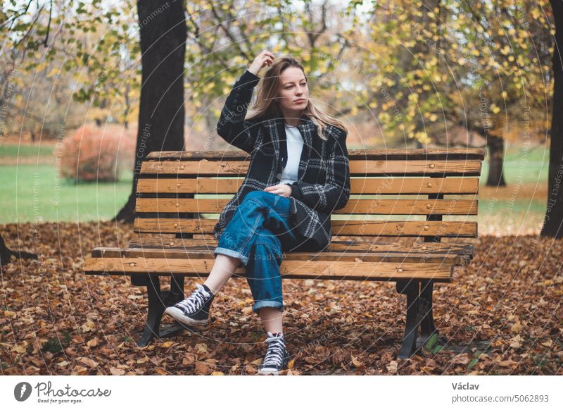 Smiling brunette sitting on an ornamental bench in the middle of the park enjoying the autumn colours. Candid portrait of a young real woman in autumn clothes