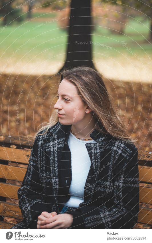 Smiling brunette sitting on an ornamental bench in the middle of the park enjoying the autumn colours. Candid portrait of a young real woman in autumn clothes