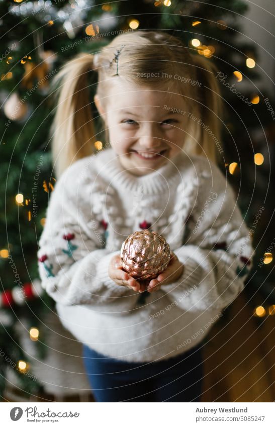 Little girl holding gold ornament in front of christmas tree Santa Claus background blonde bright celebration child childhood cute december decoration