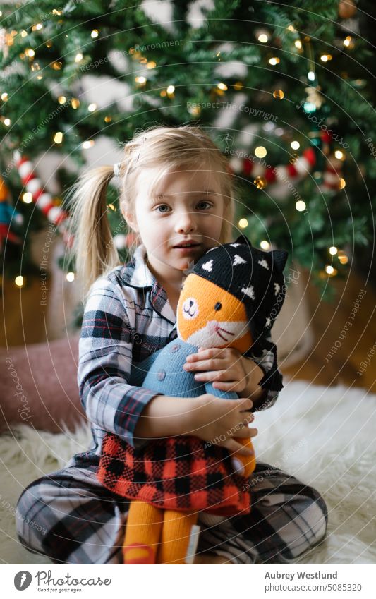 toddler girl wearing plaid pajamas cuddling with her stuffed animal in front of the Christmas tree Santa Claus background blonde bright celebration child