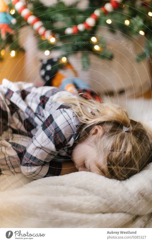 toddler girl wearing plaid pajama laying on a blanket in front of the Christmas tree Santa Claus background blonde bright celebration child childhood christmas
